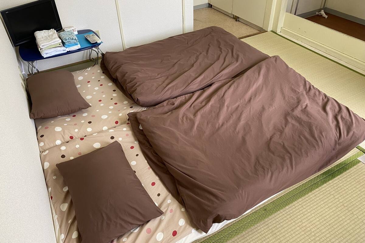 A room with two beds, each with brown bedding and a polka dot pillow. There is a small table with a flat-screen TV and a stack of folded towels.