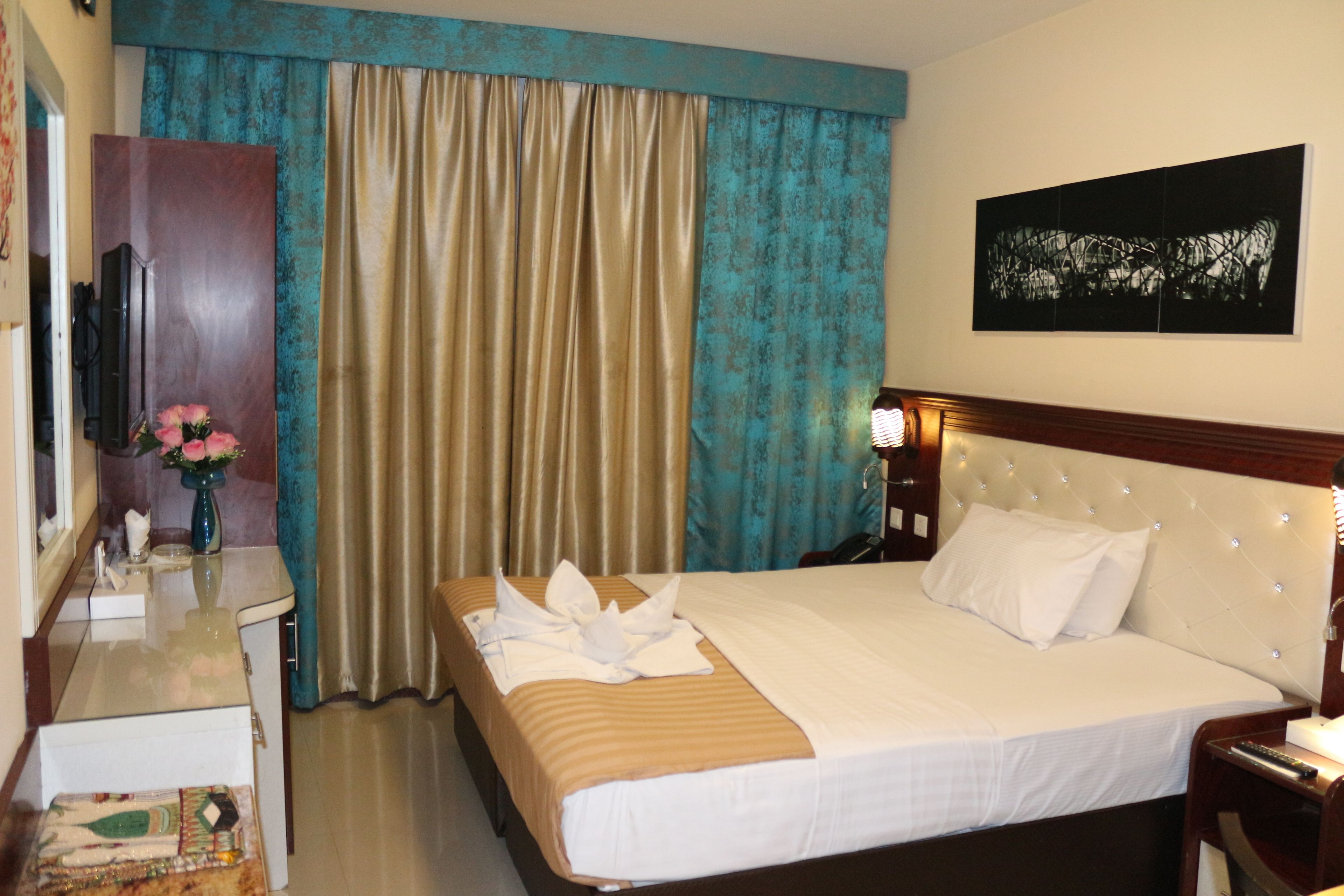 A hotel room with a bed, a desk with a vase of flowers, a dresser, and a painting on the wall.