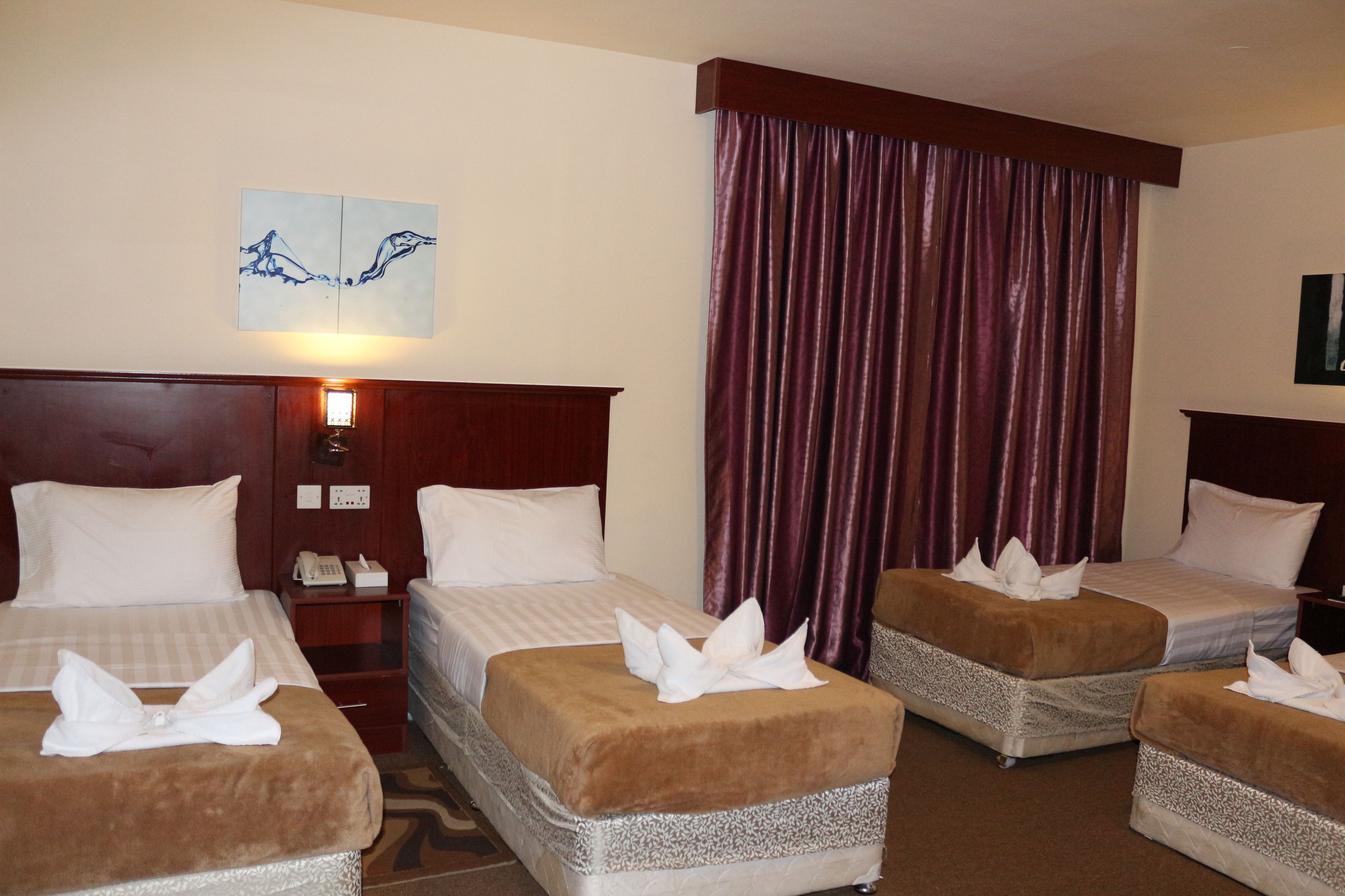 A hotel room with three beds, a large window with curtains, a wall-mounted light, and a painting.