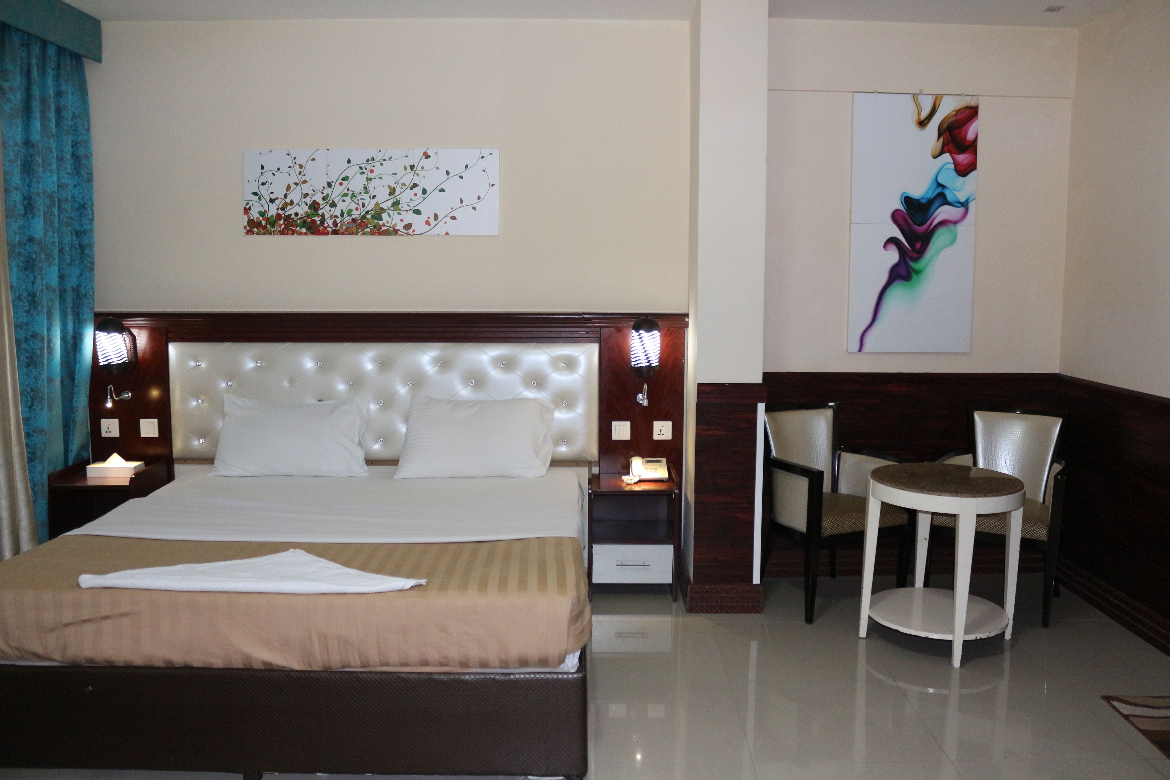 A hotel room with a large bed, a bedside table with a lamp, a chair, a small round table, and two wall art pieces.