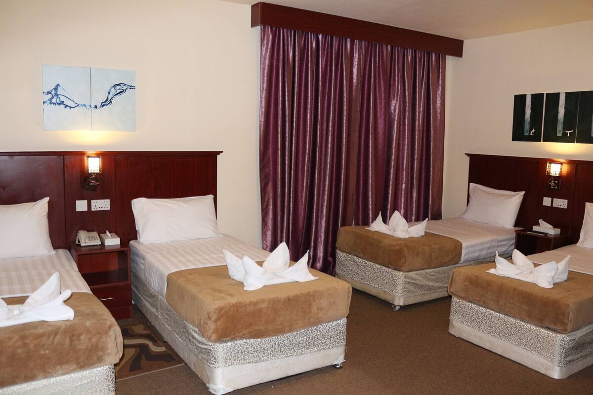 Une chambre d’hôtel avec quatre lits, chacun doté d’une tête de lit marron et de draps blancs. On y trouve deux tables de chevet avec des lampes, une grande fenêtre avec des rideaux violets et une œuvre d’art accrochée au mur.