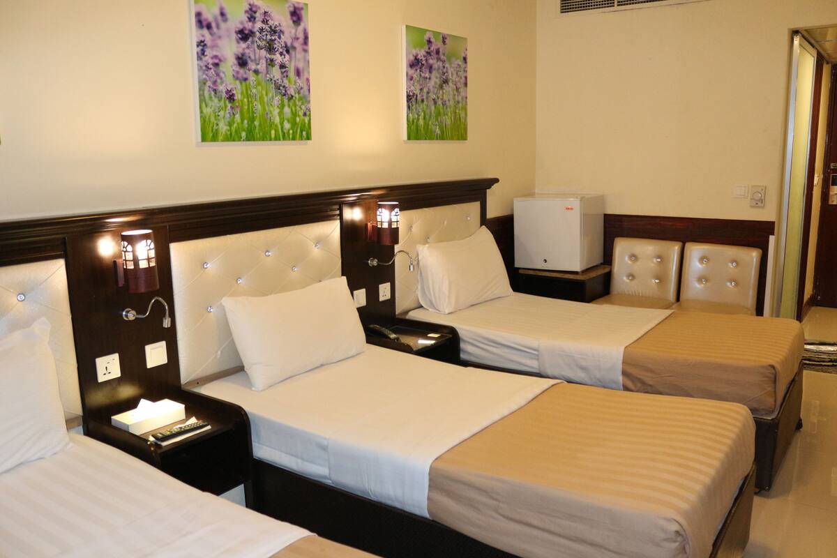 Une chambre d’hôtel avec trois lits, une table de chevet avec une lampe, une petite télévision et deux tableaux accrochés au mur.
