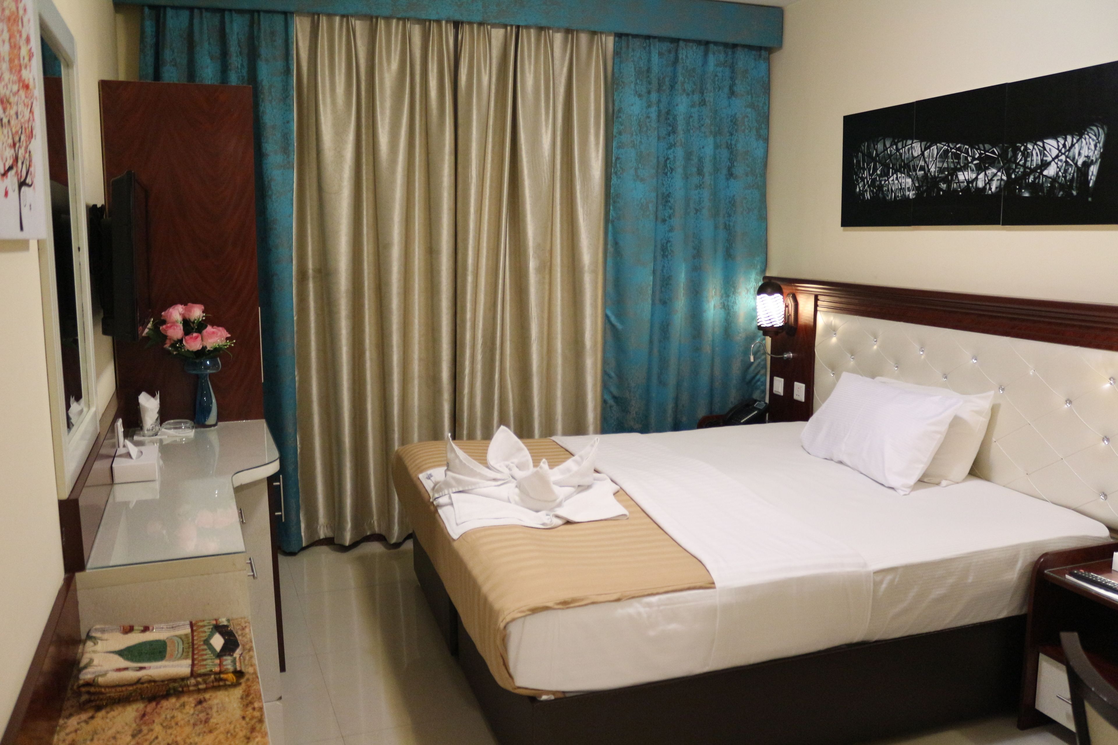 A hotel room with a bed, a bedside table, a TV, a window with curtains, and a wall-mounted artwork.