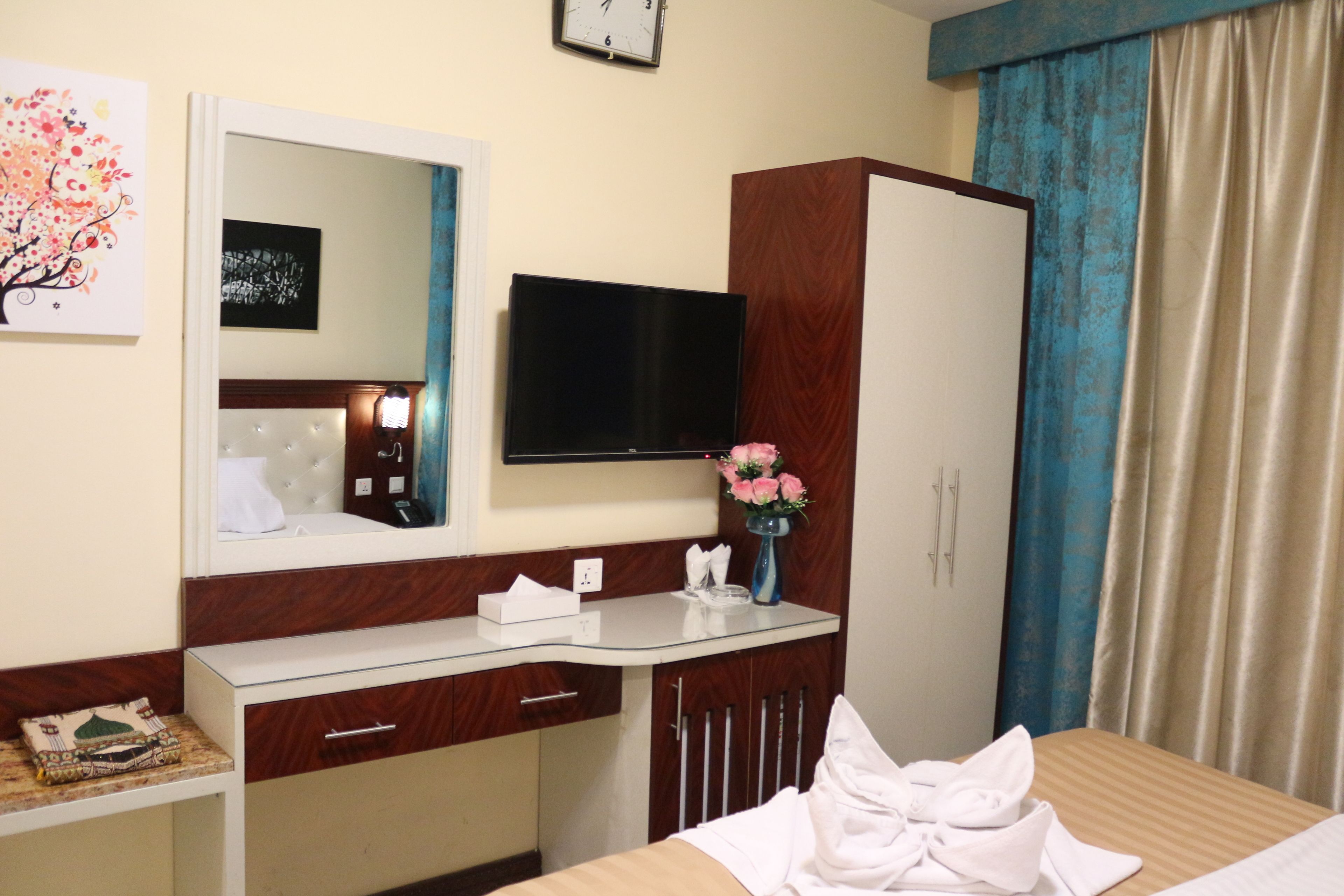 A hotel room with a bed, a vanity with a mirror, a flat-screen TV, a wooden wardrobe, a small table with a vase of flowers, and a clock on the wall.
