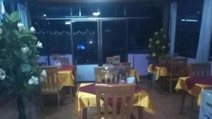 A restaurant with tables covered in yellow tablecloths, wooden chairs, and a large window with a view of the night sky. - Royal Flower Guest House (Pyin Oo Lwin)