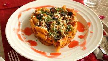 Un plato de pasta con salsa roja, decorado con aceitunas negras y hierbas, sobre un plato blanco con mantel rojo.