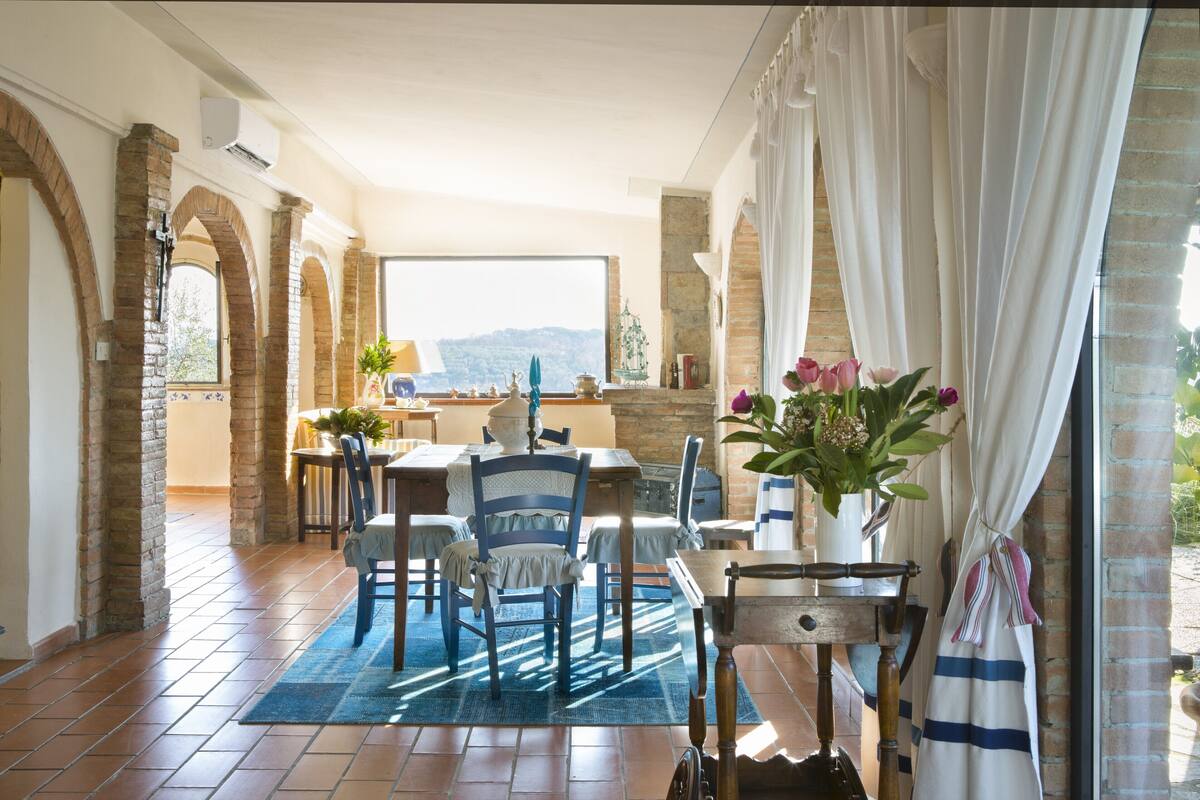A dining area with a view, featuring a table set with chairs, a blue rug, and a vase with flowers.