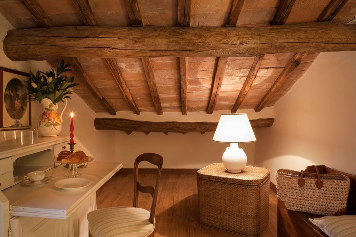 A cozy room with a wooden ceiling, a wicker table with a lamp, a chair, and a small table with a vase and a candle.