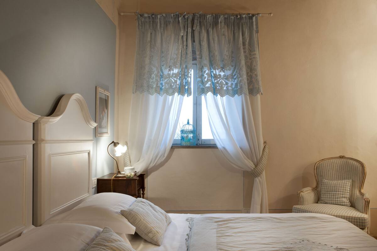 A bedroom with a large bed, a nightstand with a lamp, a chair, and a window with curtains.