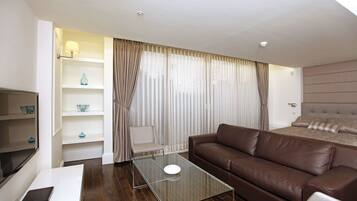 A modern hotel room with a brown leather sofa, a glass coffee table, a white TV stand, and a bed with a headboard.