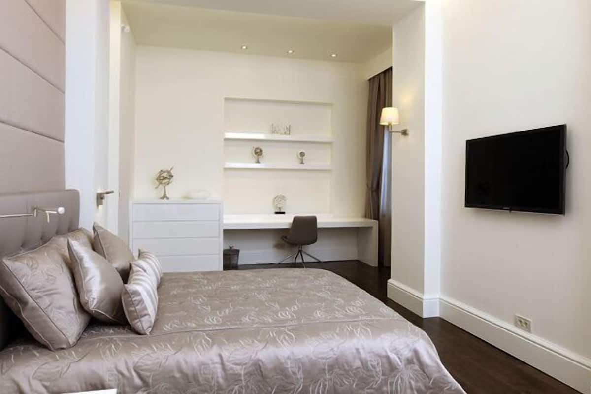 A bedroom with a bed, a desk, a chair, a TV, and a shelf with decorative items.