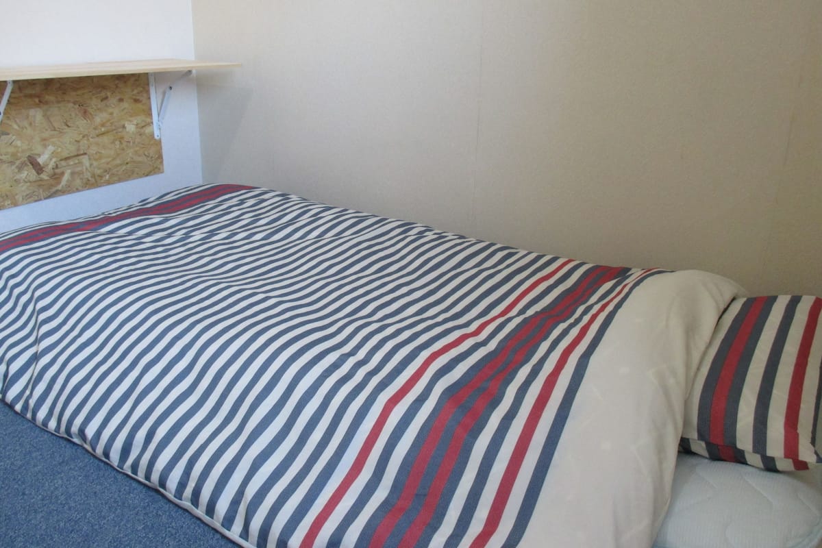 A single bed with a striped bedspread and pillows, a wooden headboard, and a plain wall.
