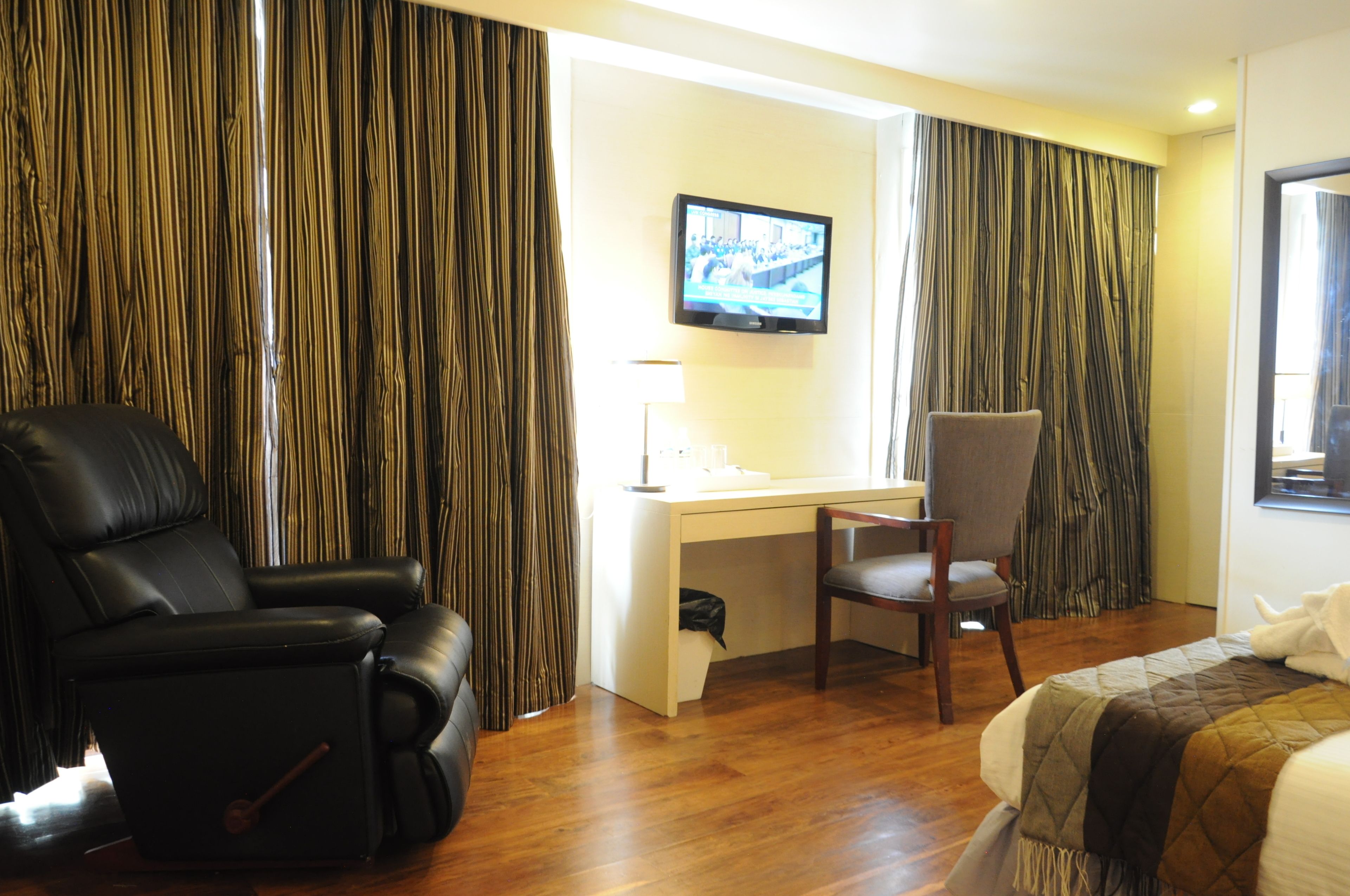 Ein Hotelzimmer mit schwarzem Ledersessel, Schreibtisch inklusive Computer, Wand-TV und einem Bett mit gesteppter Decke.