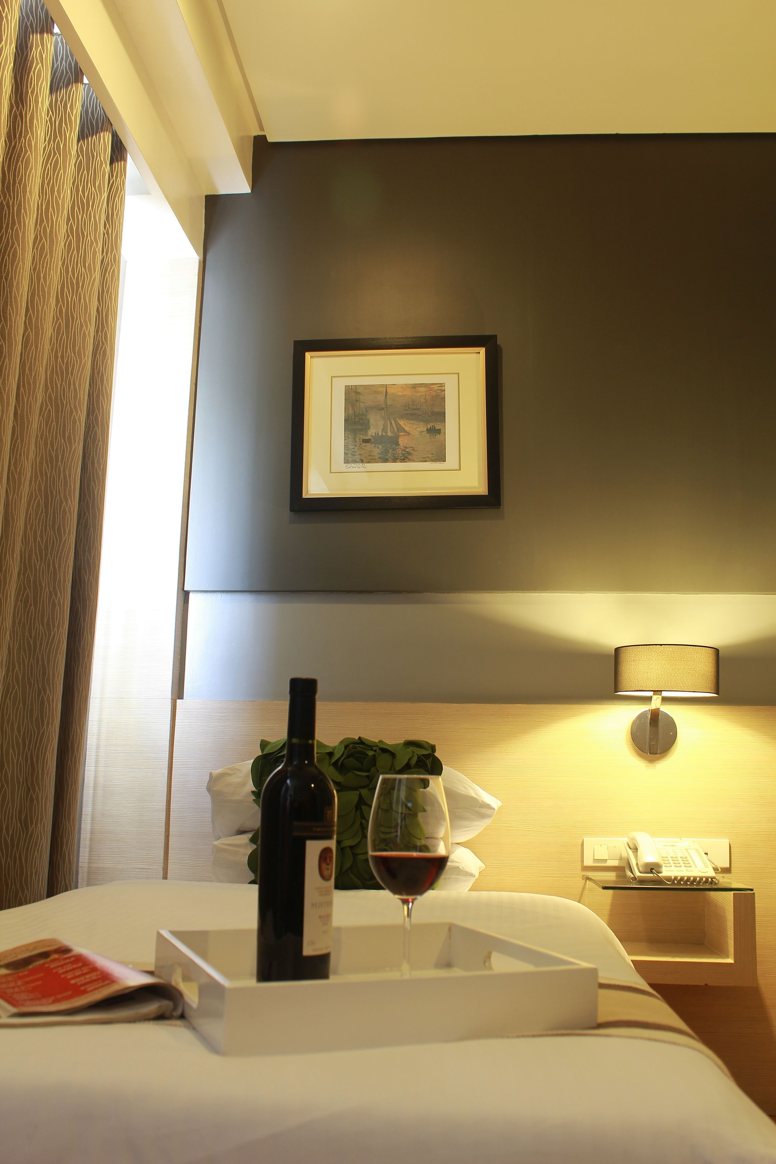 Quarto de hotel com uma cama, mesa de cabeceira com uma garrafa de vinho e um copo, um quadro na parede e uma luminária.