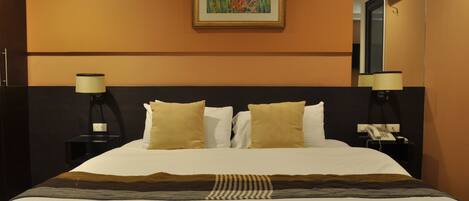 Quarto de hotel com uma cama, duas luminárias de cabeceira, um quadro na parede e um telefone em uma mesinha de cabeceira.