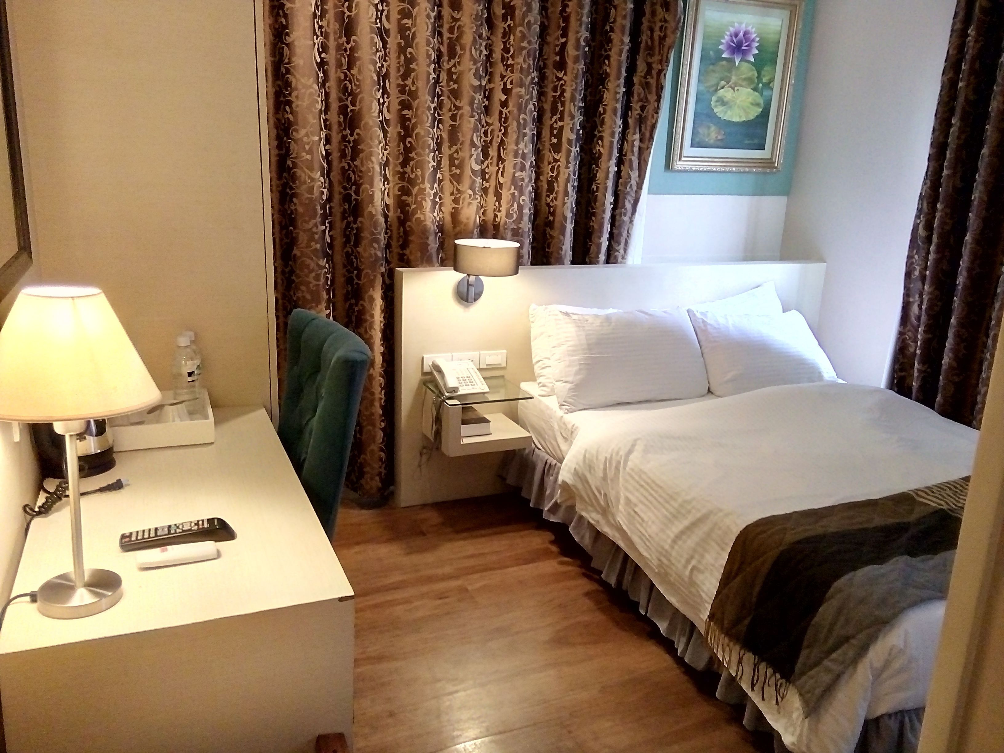 Une chambre d’hôtel avec un lit, un bureau avec une lampe, une chaise, une table de chevet avec un téléphone et des rideaux.