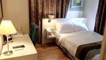 Une chambre d’hôtel avec un lit, un bureau avec une lampe, une chaise, une table de chevet avec un téléphone et des rideaux.