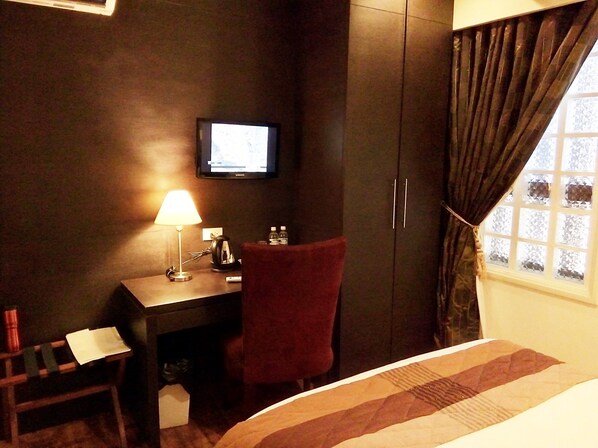A hotel room with a bed, a desk with a lamp, a chair, a TV on the wall, and a wardrobe. - Ciudad Fernandina Hotel (Vigan)