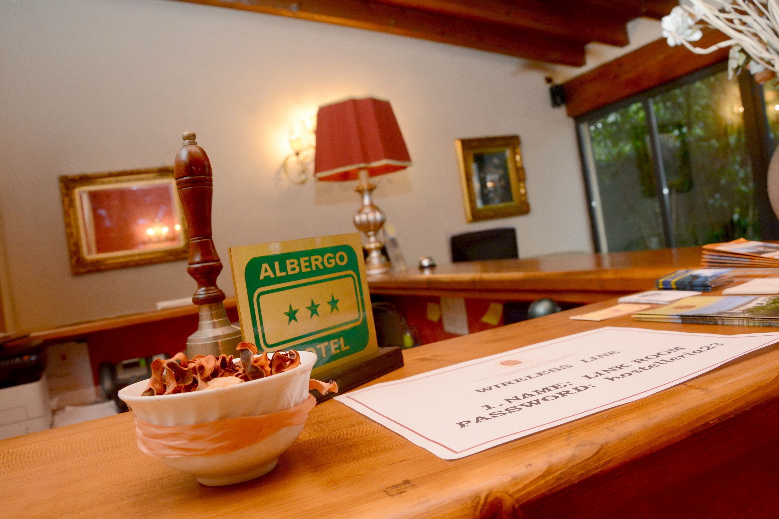 Bancada de recepção de hotel com uma placa indicando "Albergo Hotel", uma tigela de lanches, uma luminária e uma janela com vista para áreas verdes.