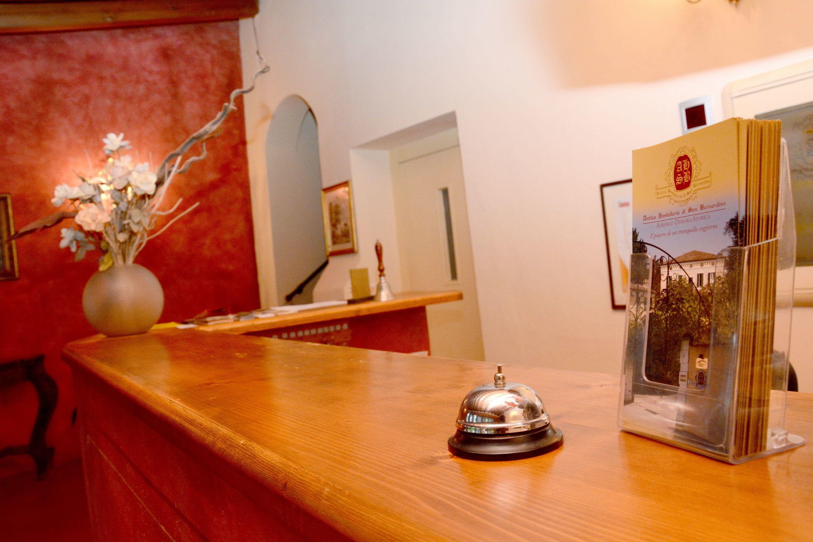 A reception desk with a wooden counter, a metal bell, a vase with flowers, and a framed picture on the wall.