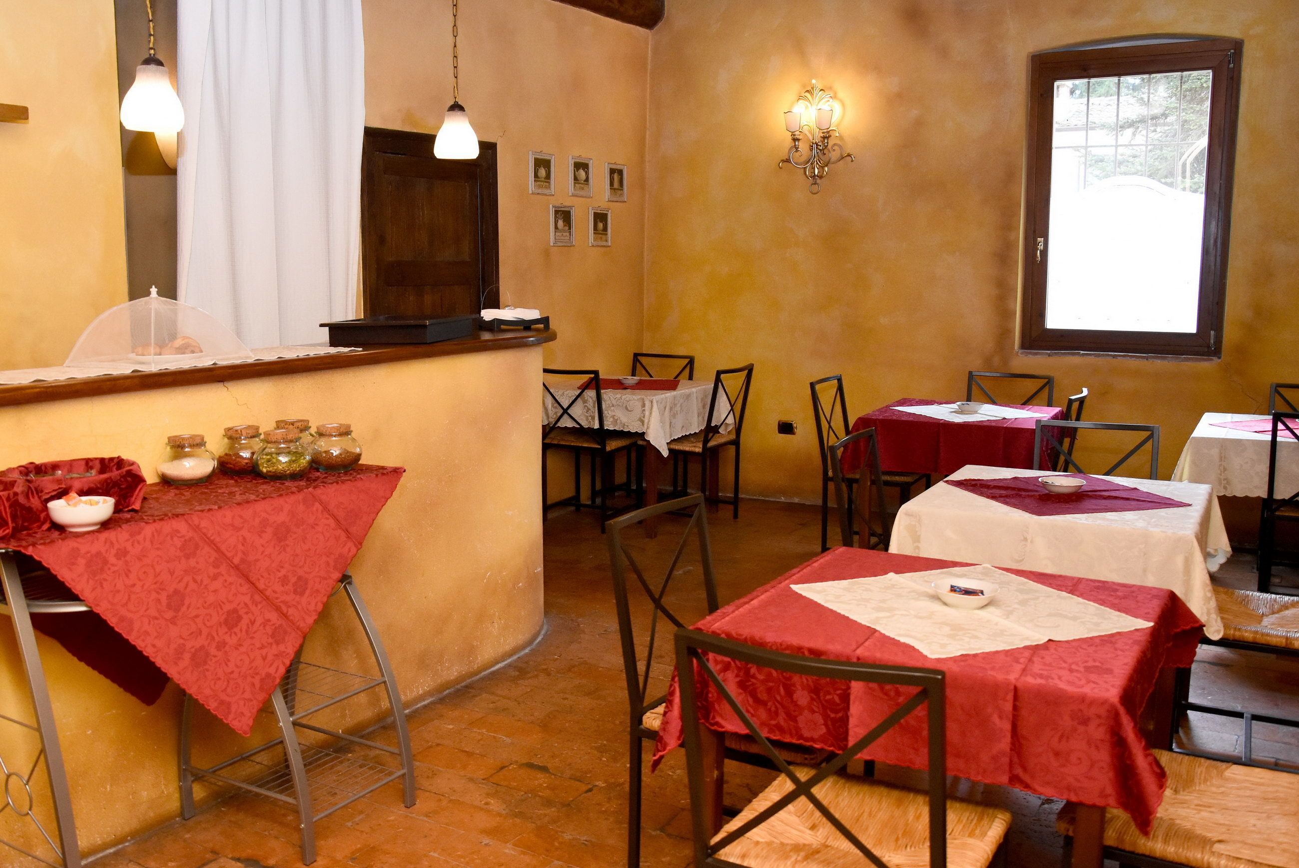 A restaurant with tables covered in red tablecloths, a bar area with jars of condiments, and a window with a view of the outside.