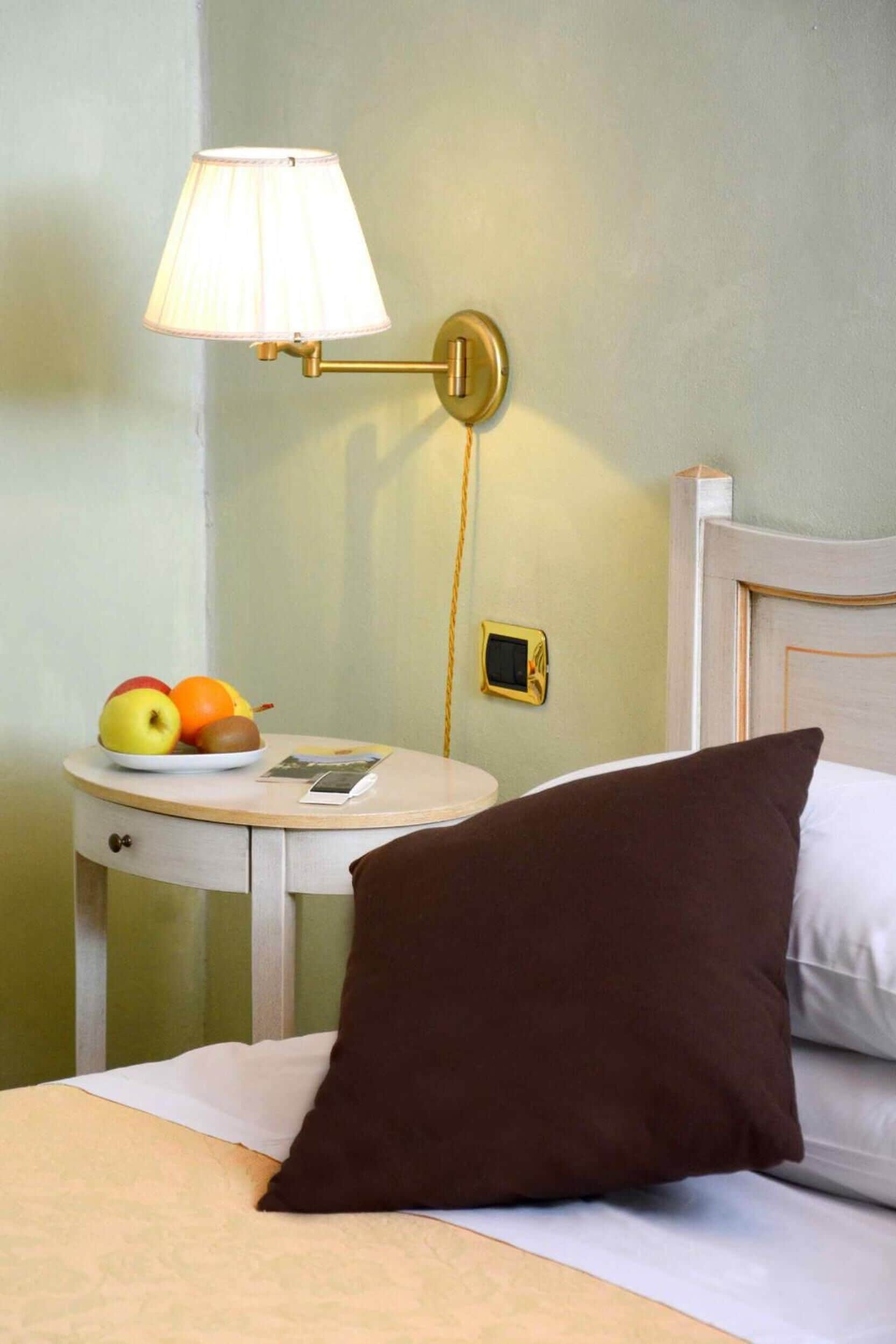 Ein Hotelzimmer mit einem Bett, einem Nachttisch mit Lampe, einer Obstplatte und einer kleinen digitalen Uhr.