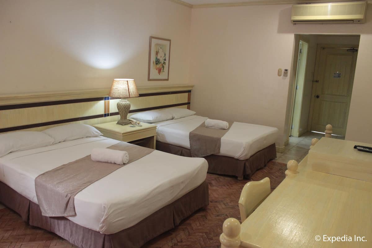 Habitación de hotel con dos camas individuales, una mesita de noche con lámpara, una pequeña mesa con sillas y una puerta que da a otra habitación.