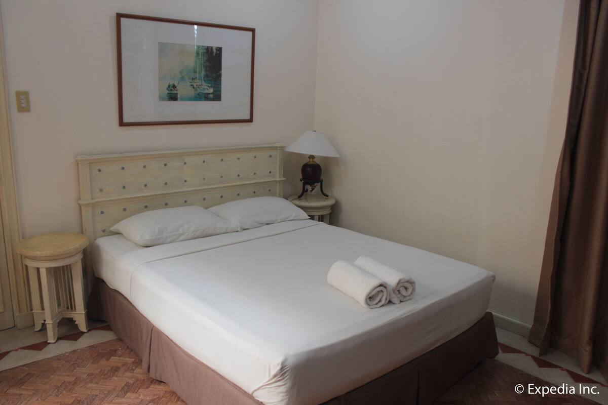 Una cama bien hecha con sábanas blancas y una toalla enrollada al lado, una lámpara de noche y un cuadro en la pared.