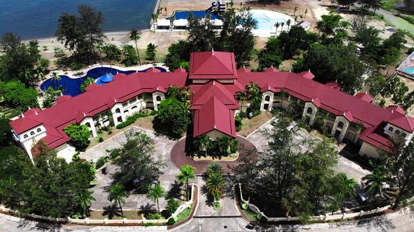 An aerial view of a resort with a central building, multiple guest rooms, a swimming pool, and surrounding greenery.