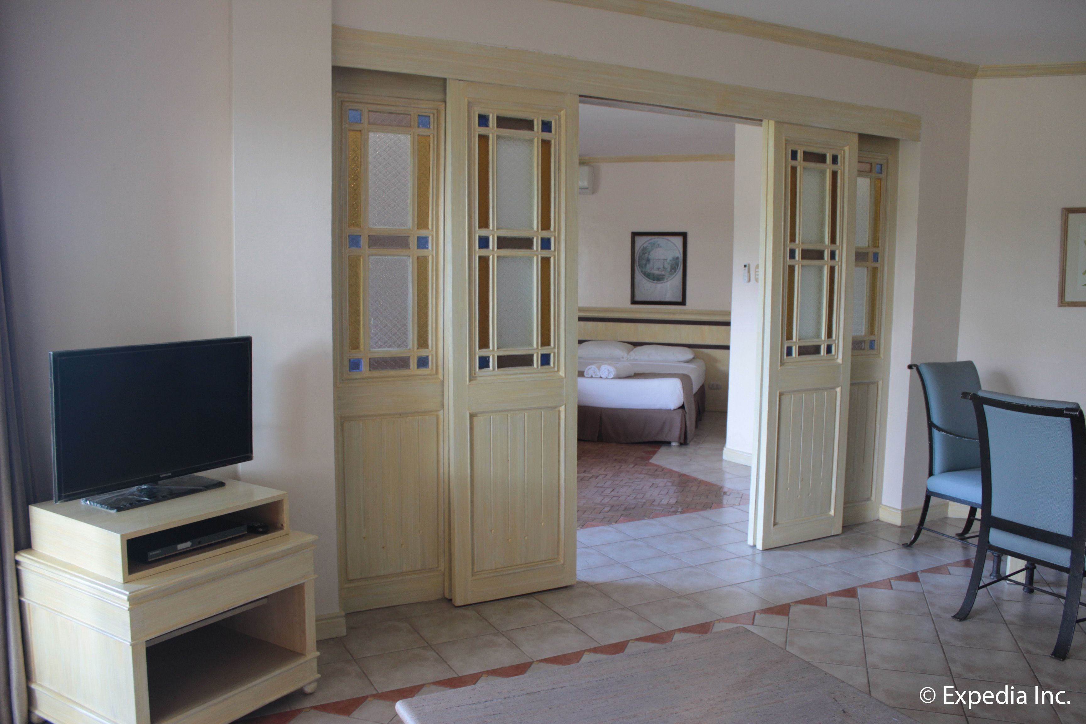 Habitación de hotel con televisión sobre un soporte de madera, cama con cabecera blanca, una silla y una puerta que da a otra habitación.