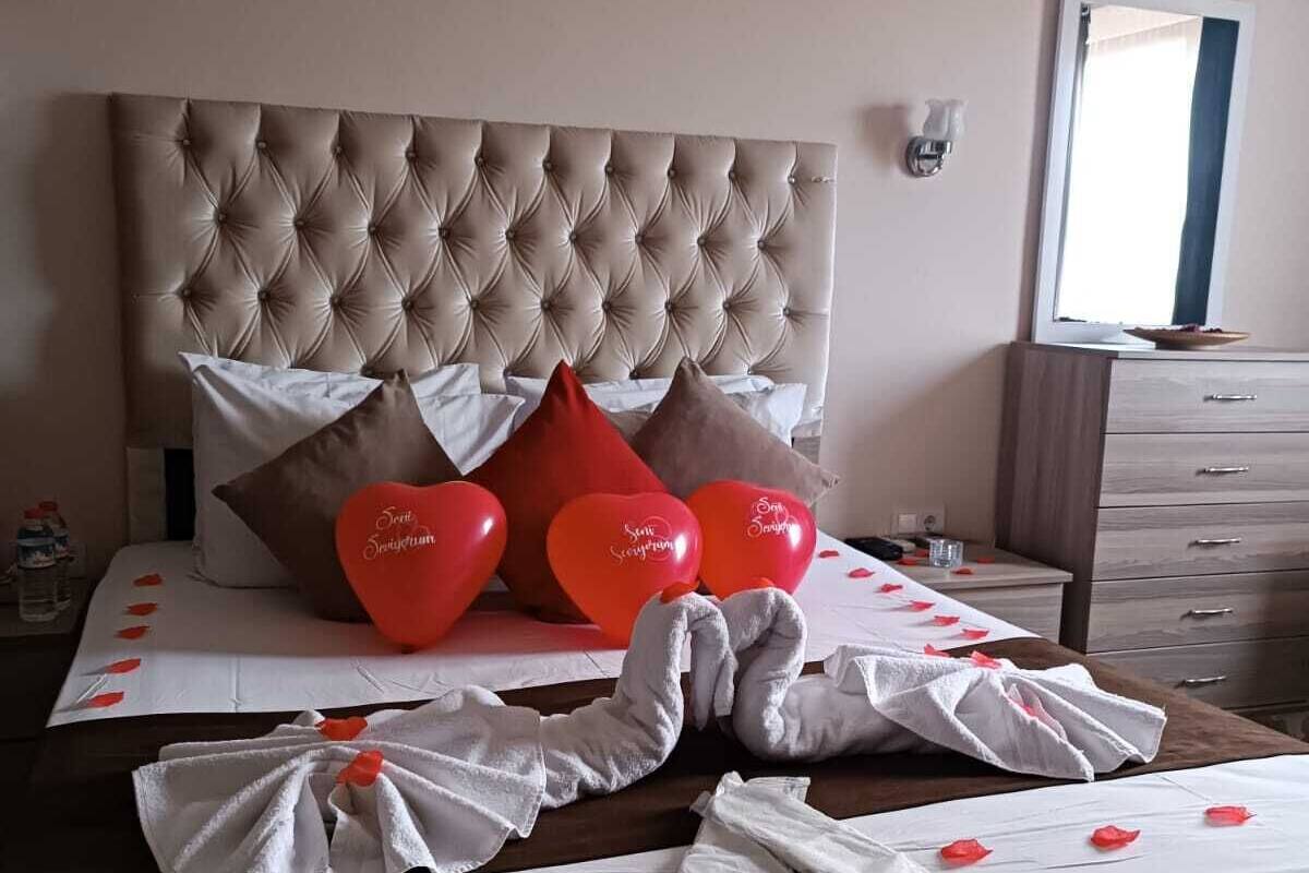 Quarto de hotel com cabeceira estofada, cama com roupa de cama branca e almofadas em formato de coração vermelho, e mesa de cabeceira com uma garrafa e copos.