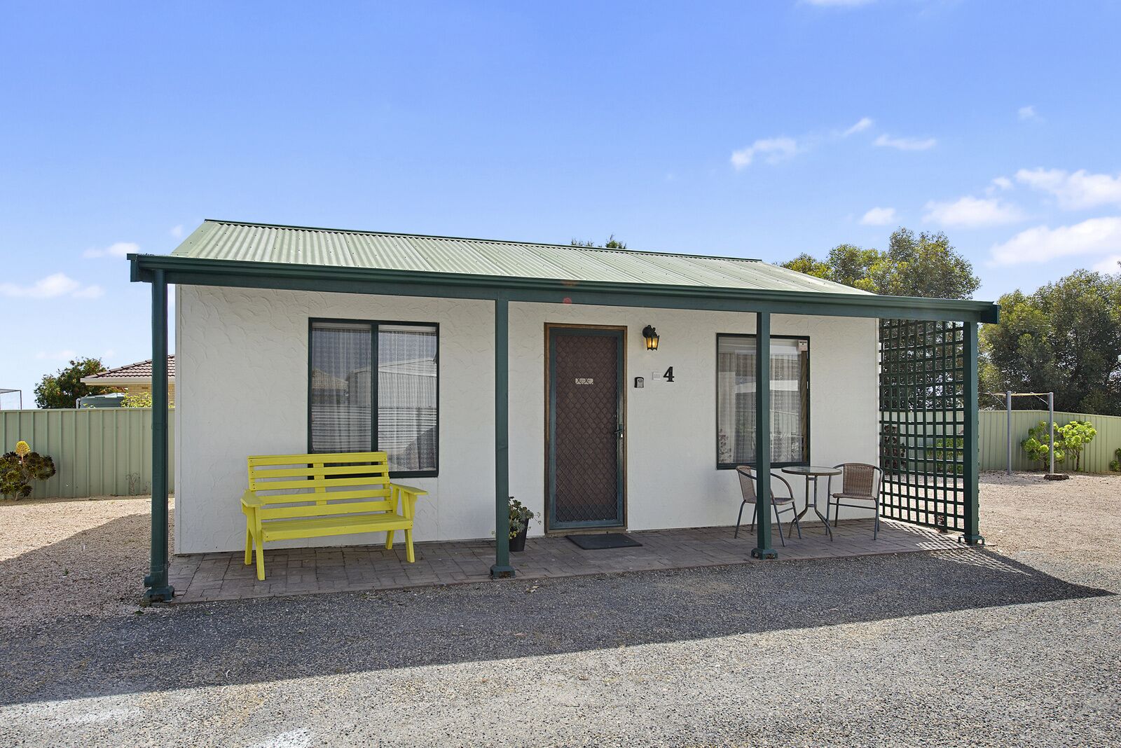 A small, single-story building with a green roof and white walls. It features a bench, a door, and a small table with chairs.