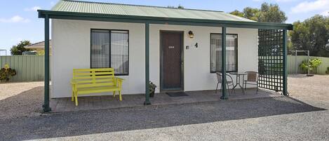A small, single-story building with a green roof and white walls. It features a bench, a door, and a small table with chairs.