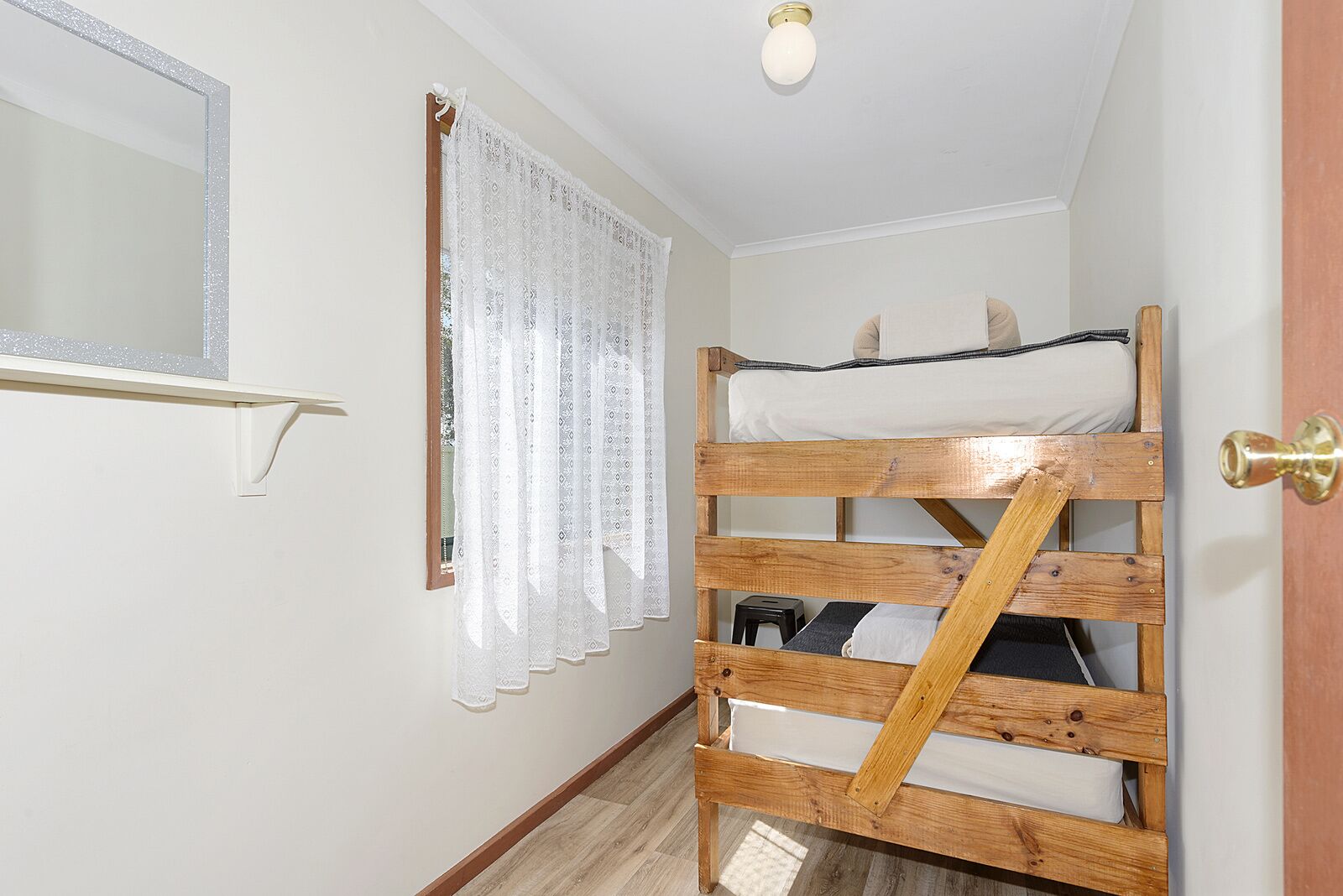 Una habitación pequeña y funcional con una litera, un espejo, una ventana con cortinas de encaje y una puerta con tirador de latón.