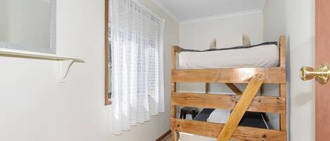 Una habitación pequeña y funcional con una litera, un espejo, una ventana con cortinas de encaje y una puerta con tirador de latón.