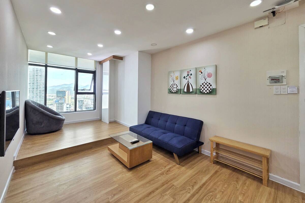 A modern living room with a blue sofa, a glass coffee table, and a wooden bench. There is a large window with a city view, and the room has wooden flooring.
