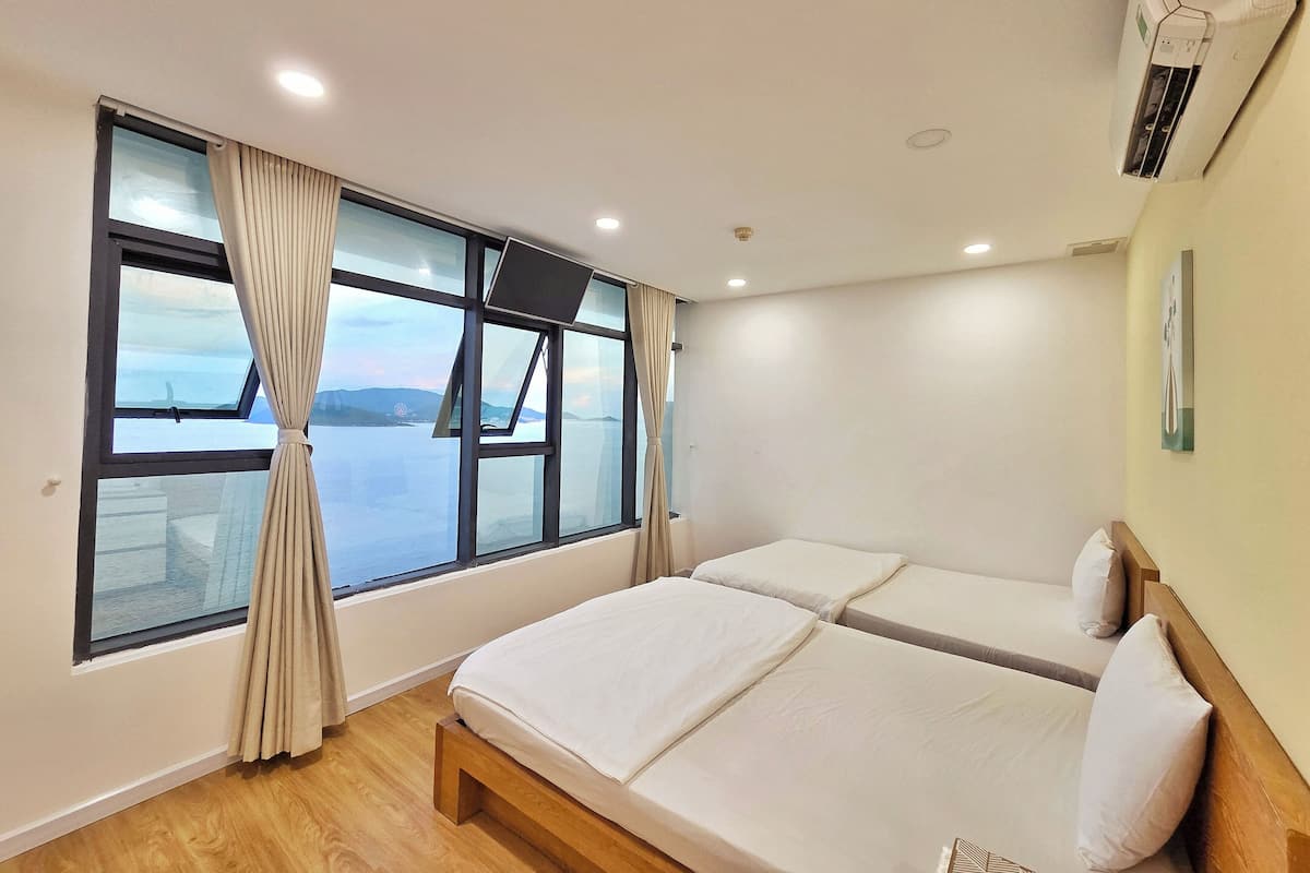 A hotel room with two beds, a large window with curtains, a wooden floor, and an air conditioning unit on the wall.
