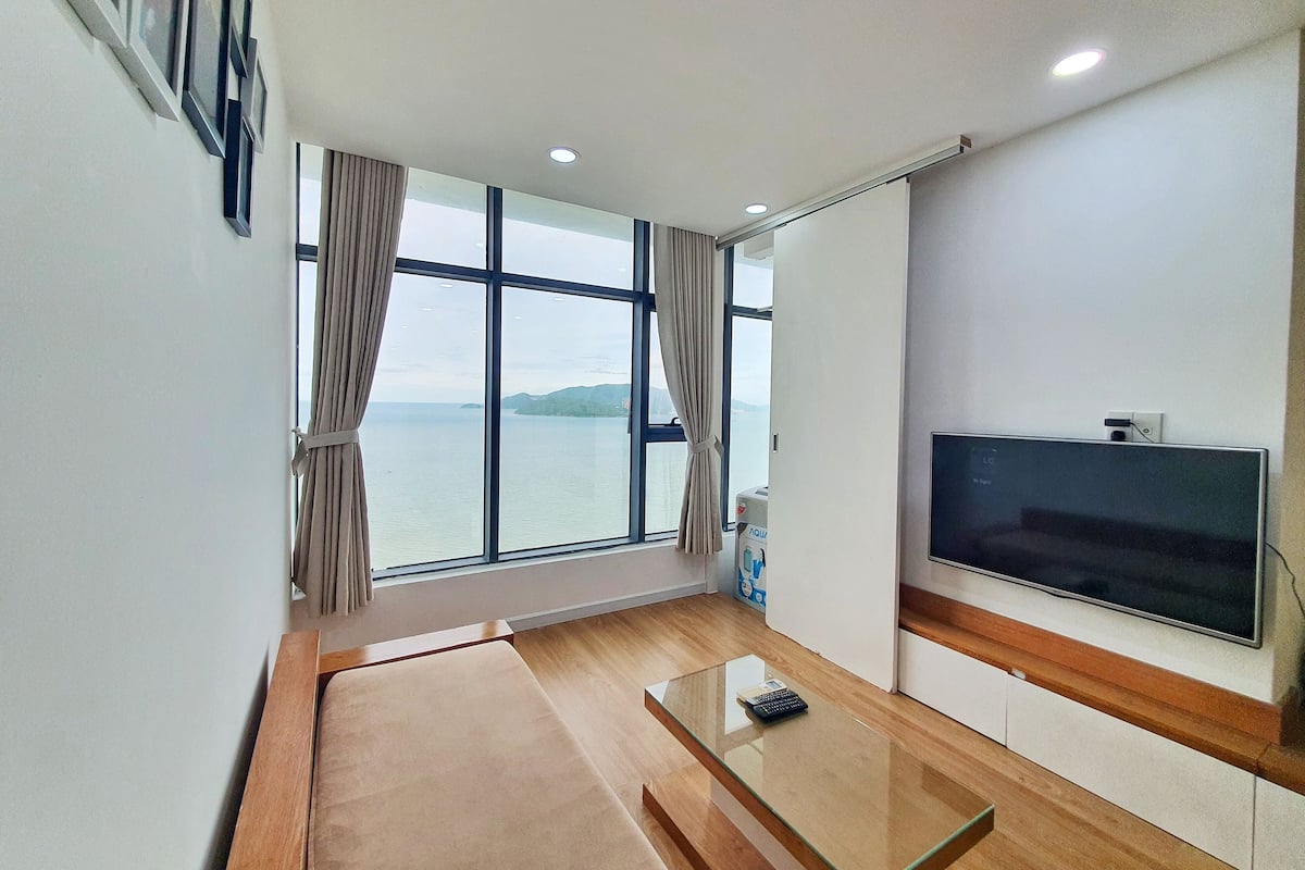 A modern living room with a large window, a flat-screen TV, and a glass coffee table.
