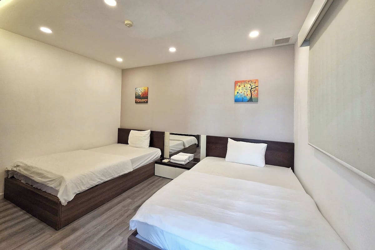 A hotel room with two single beds, a wooden headboard, a small bedside table with a lamp, and a wall-mounted artwork.