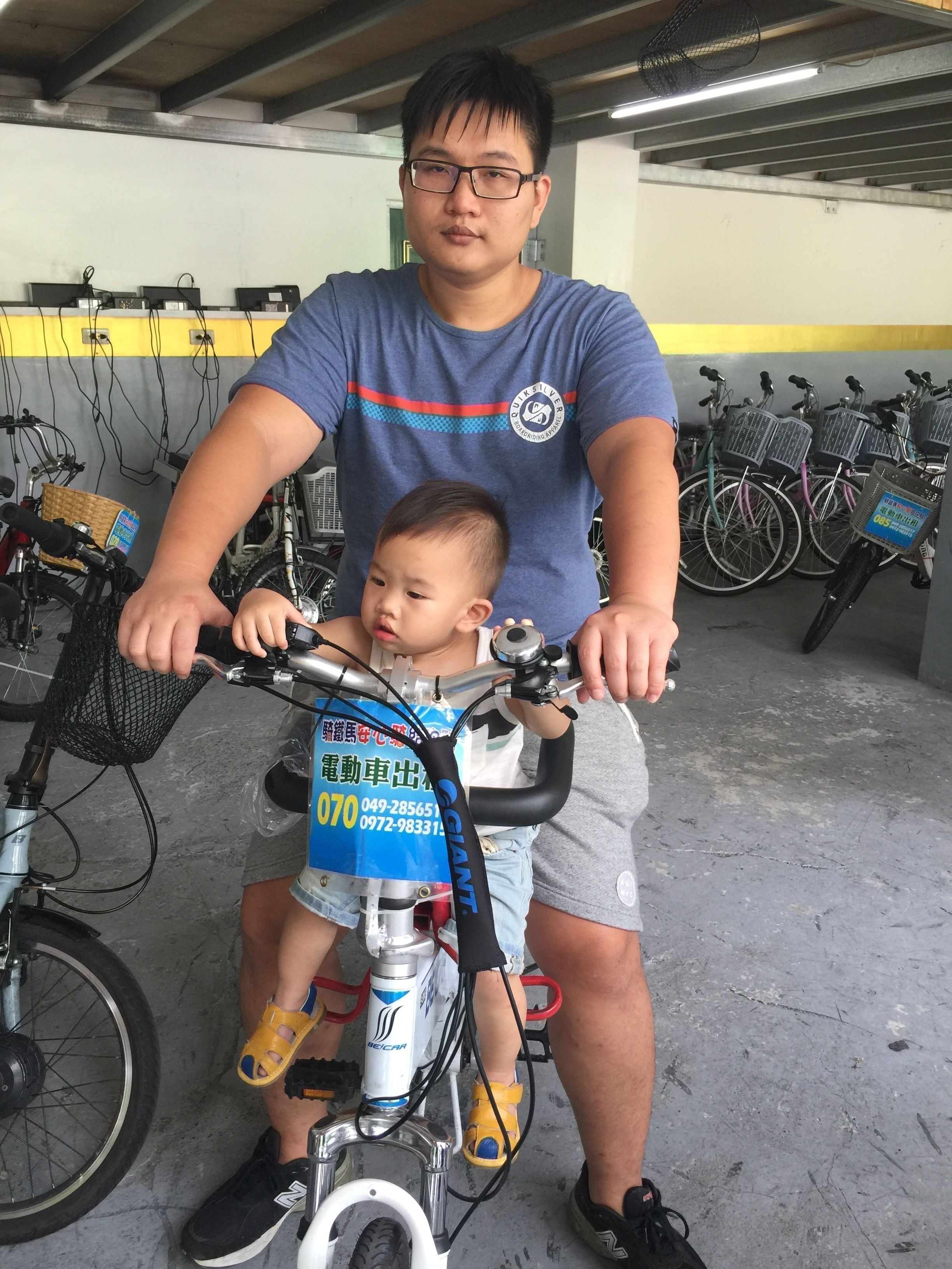 A man and a child on a bicycle in a parking garage with bicycles and a basket in the background.
