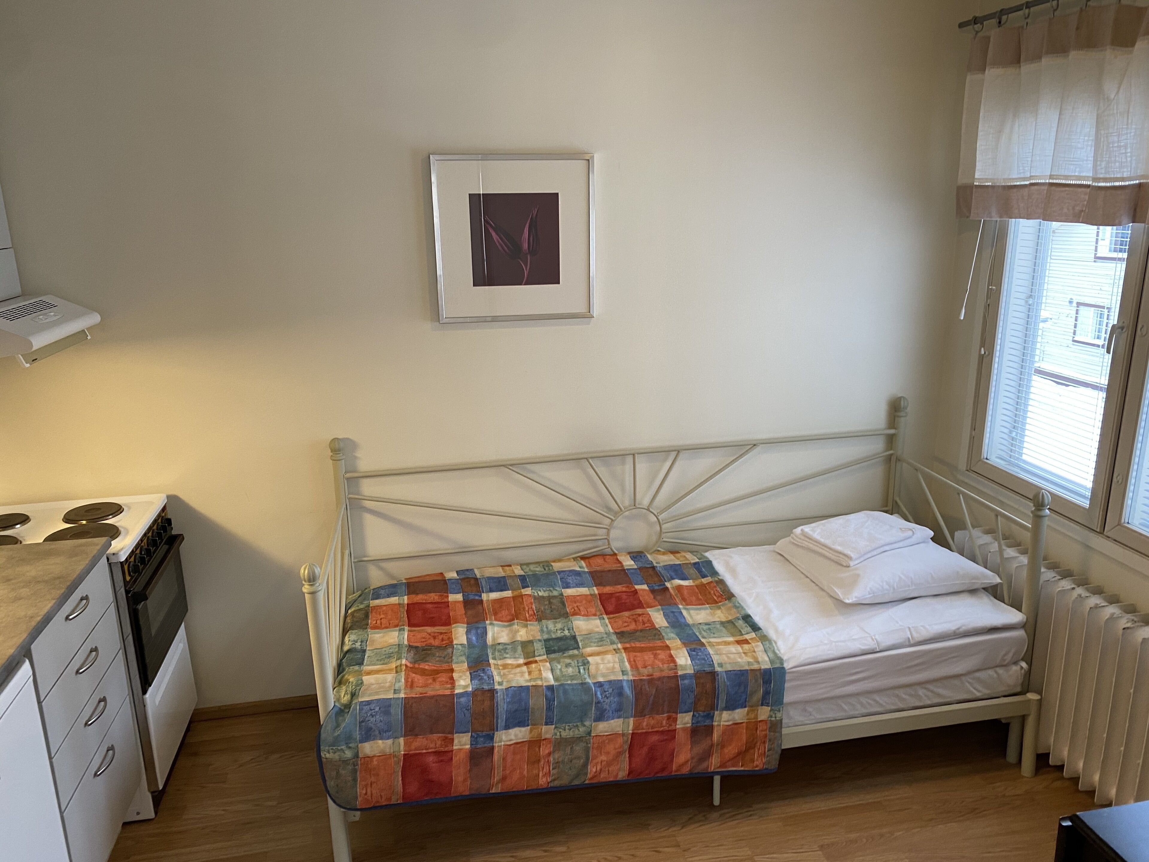 A small, single-bed room with a metal headboard, a window with curtains, a radiator, and a kitchenette with a stove and oven.