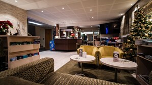 Eine Hotelhalle mit einer Rezeption, zwei gelben Sesseln, einem Couchtisch, einem Weihnachtsbaum und einem Bücherregal mit Büchern.