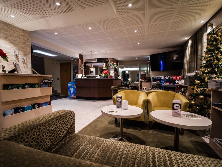 Un hall de hotel con mostrador de recepción, dos sillones amarillos, una mesa de centro, un árbol de Navidad y una estantería con libros.