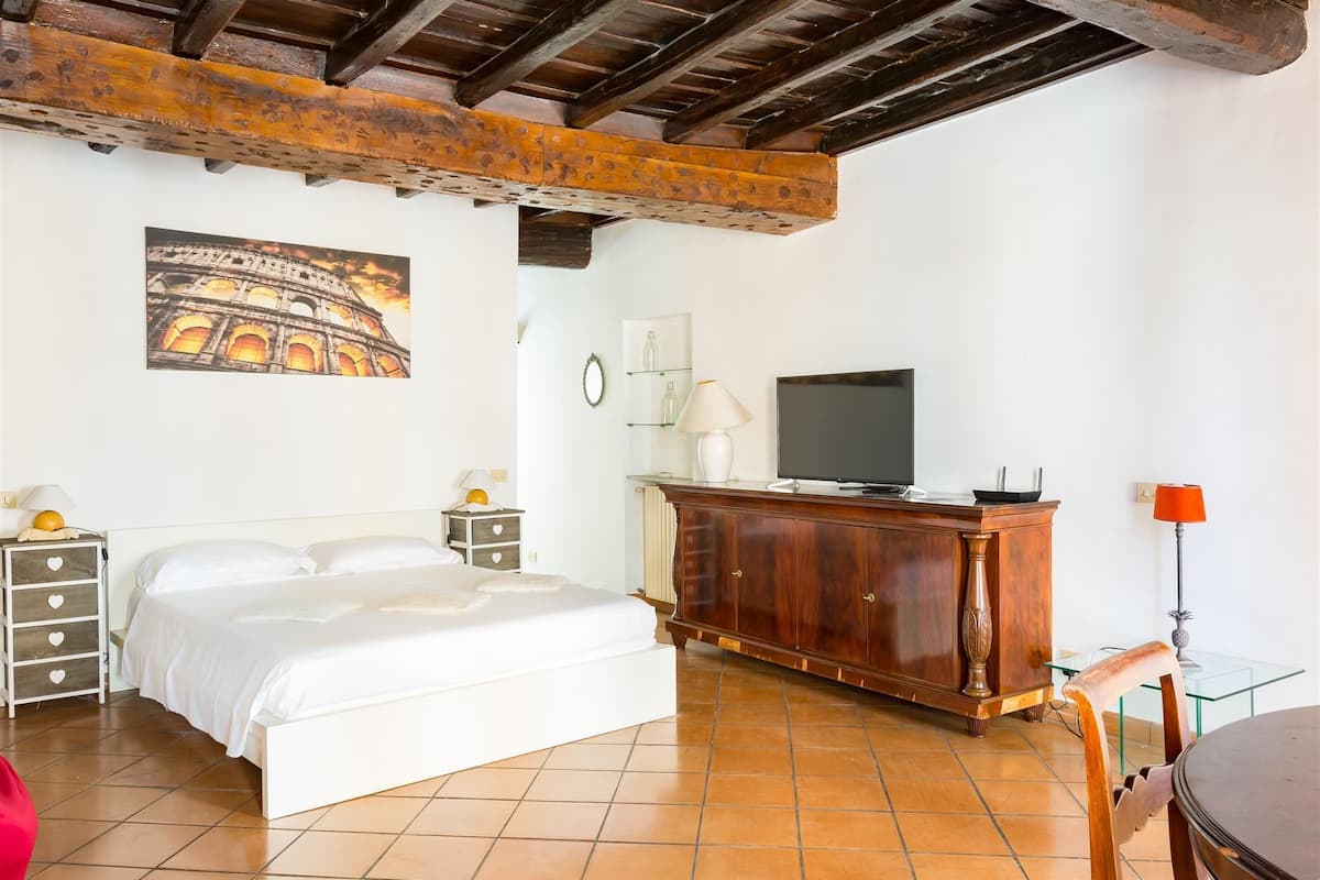 A bedroom with a bed, wooden ceiling beams, a TV, a dresser, and a glass table.