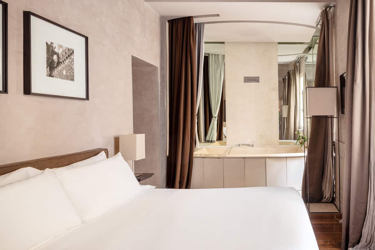 A hotel room with a bed, a bedside table with a lamp, a large mirror, and a bathroom visible through an open door.