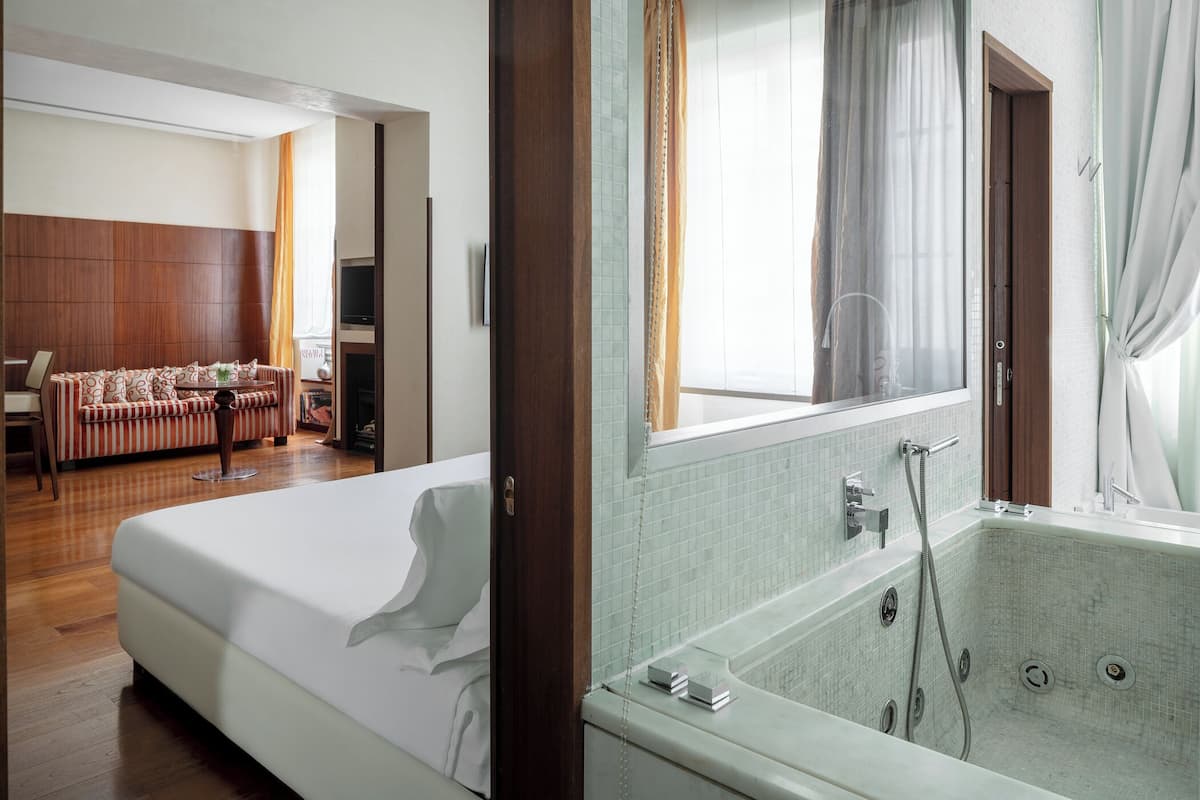 A hotel room with a bed, a sofa, a television, a bathroom with a bathtub, and a window with curtains.