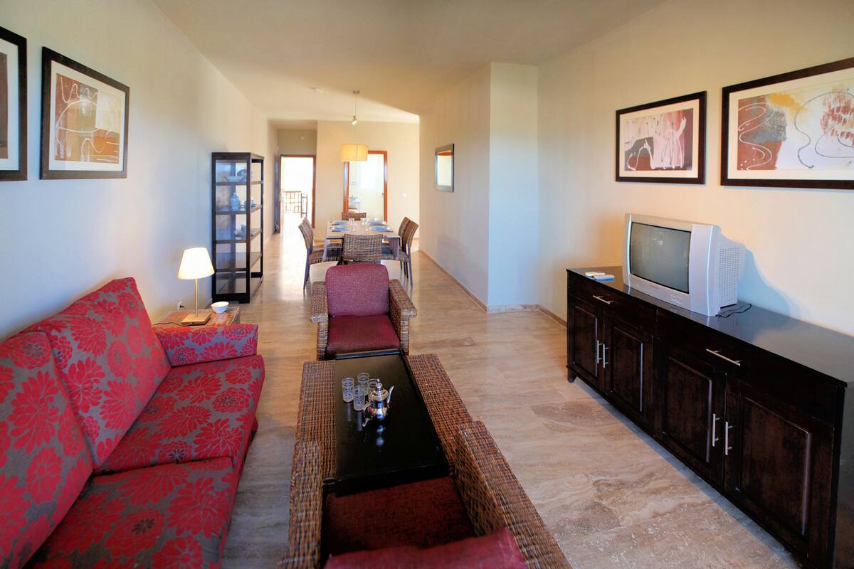 A living room with a red sofa, a coffee table, a TV, and a dining area in the background.