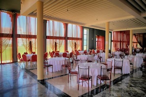 A spacious banquet hall with multiple round tables set for dining, large windows with sheer curtains, and a polished marble floor. - La Perle Orientale - Saïdia (Madagh)