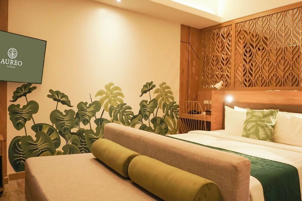 A hotel room with a bed, a wooden headboard, a TV mounted on the wall, and a decorative plant wall.