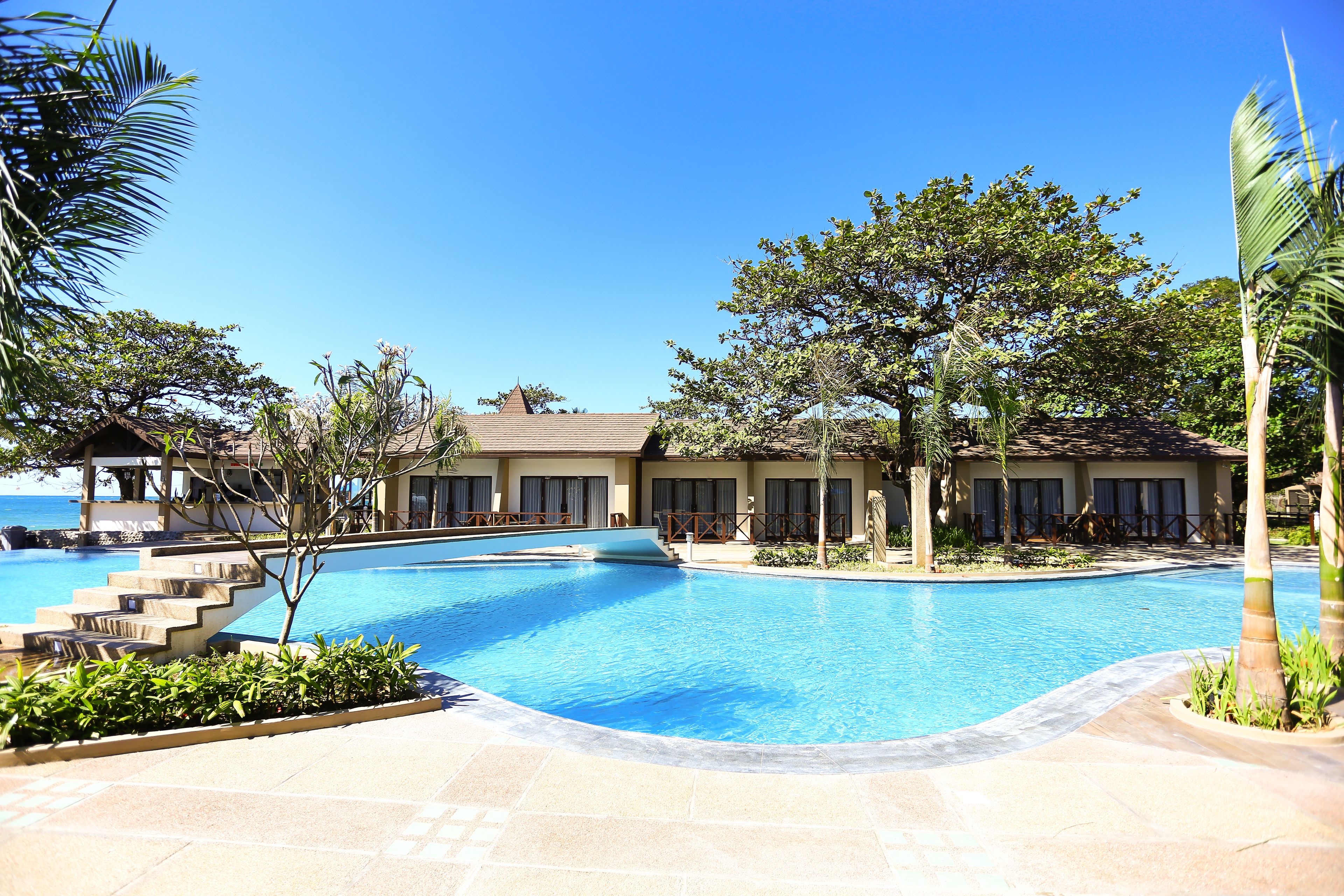 Piscina com degraus de acesso, rodeada por palmeiras e um edifício com telhado de azulejos.
